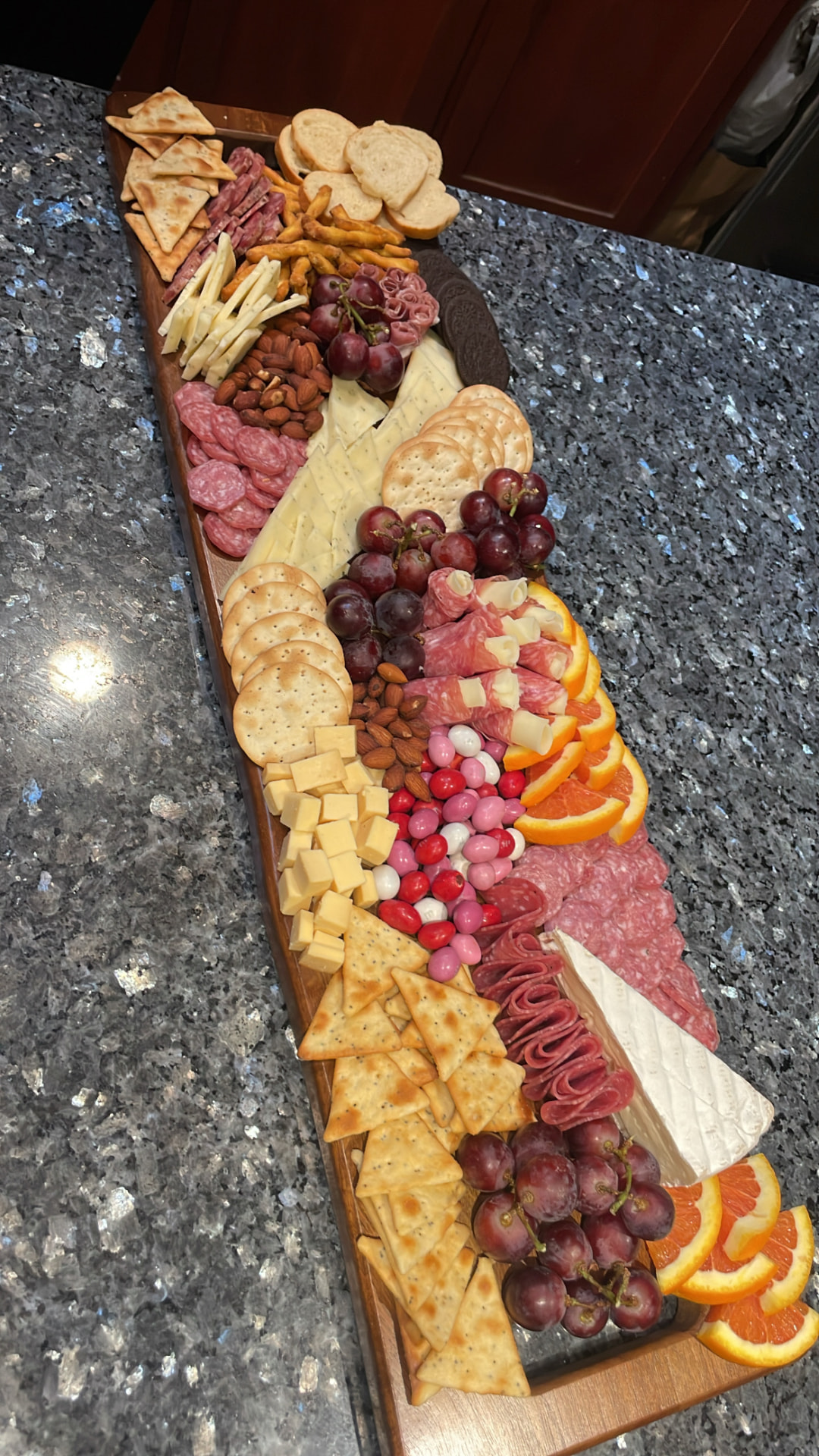 Long wooden grazing board with crackers, salami, cheeses, grapes, orange slices and candies